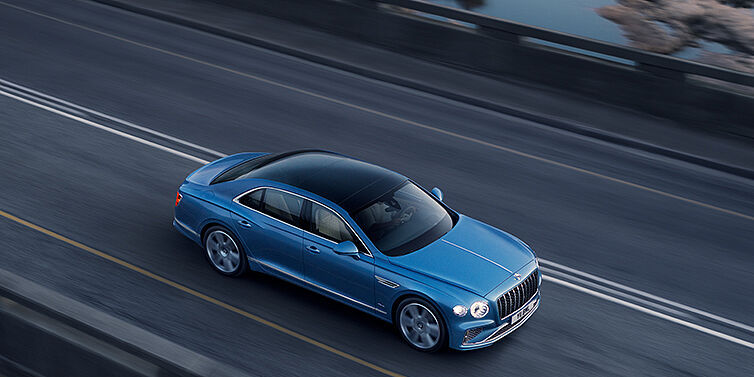 Bentley Cyprus New Bentley Flying Spur Azure sedan in Light windsor blue paint by Mulliner, with 22 inch Azure wheels seen from overhead driving dynamically on a country road