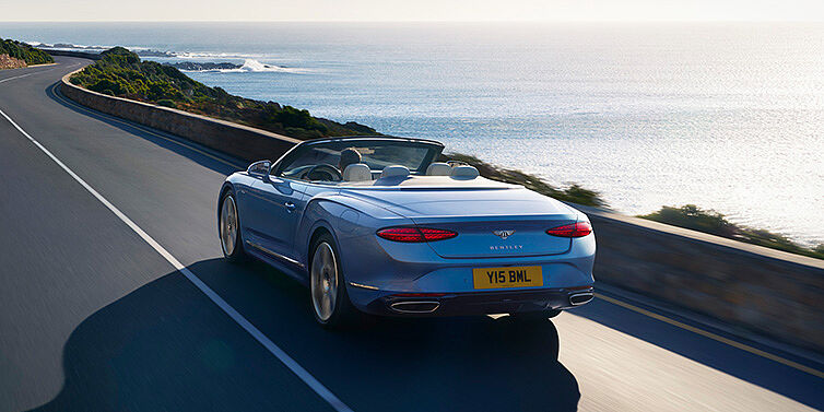 Bentley Cyprus New Bentley Continental GTC Azure convertible rear view in Light Windsor blue paint with 22 inch ten spoke wheels driving dynamically on an ocean road in the sunshine